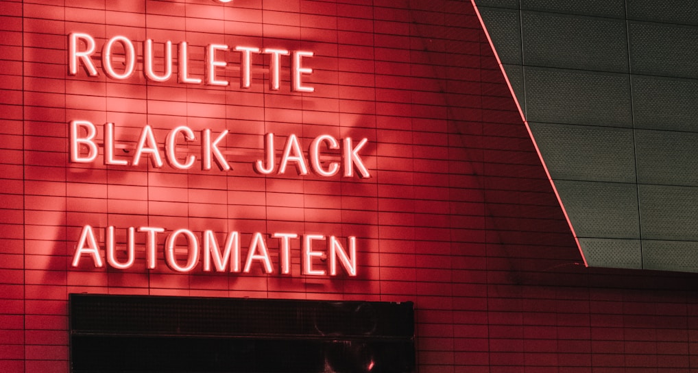 Red neon letters are mounted on a dark tiled wall, displaying the words Poker, Sic Bo, Roulette, Black Jack, and Automaten.