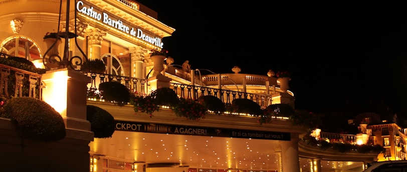 A luxurious casino at night, illuminated with warm, golden lights. The building features elegant architecture with columns and decorative elements. A few people are walking towards the entrance, and there are a couple of parked cars nearby.