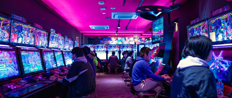 A vibrant arcade with rows of gaming machines adorned with colorful screens. Several people are immersed in playing games, seated on stools in a dimly lit room accented by an intense pink ceiling light. The atmosphere is lively and engaging.