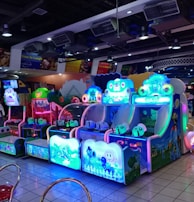 A brightly lit arcade filled with colorful gaming machines sits under ceiling lights. Neon lights illuminate various game stations, creating a playful and vibrant environment. A few red chairs are scattered around the reflective, tiled floor, and a person is observing one of the machines. The atmosphere is lively and cheerful, featuring games with cartoonish designs and engaging themes.